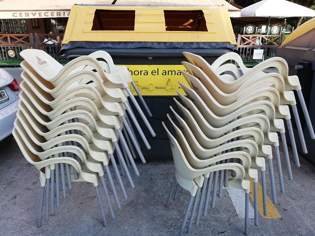 Sillas de una terraza de hostelería apiladas junto a un contenedor de reciclaje.